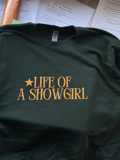 Green sweatshirt with 'Life of a Showgirl' text on a table with papers underneath